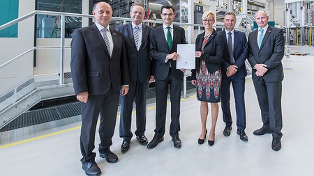 Six persons can be seen. One man and one woman in the middle are holding a sheet of paper.