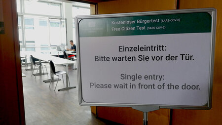 Ein Schild steht vor einem Raum, dessen Tür geöffnet ist. Im Raum sitzt ein Mann an einem Tisch.