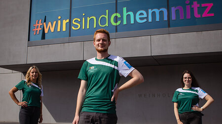 Ein junger Mann und zwei kunge Frauen in Laufshirts der TU Chemnitz stehen vor dem Weinholdbau. Im Hintergrund ist der Hashtag wirsindchemnitz zu sehen. 