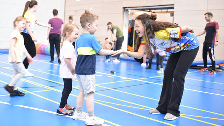 Eine junge Frau leitet Kinder in euiner Sporthalle beim Turnen an.